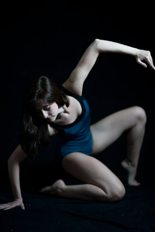 Dancer kneels in black One leg and arm raised and bent Wears blue leotard
