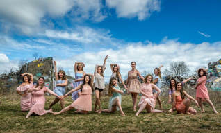 Fourteen dancers pose in two lines outside in front of a blue sky with white clouds. 