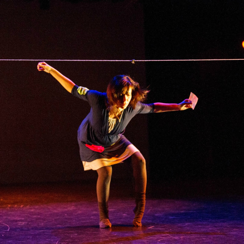 Dancer in black room Arms out to side, holds a note Clothesline in background