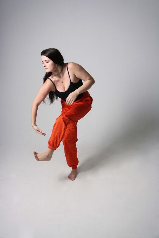 Dancer against all white In black tank top and red pants Reaches arm to foot