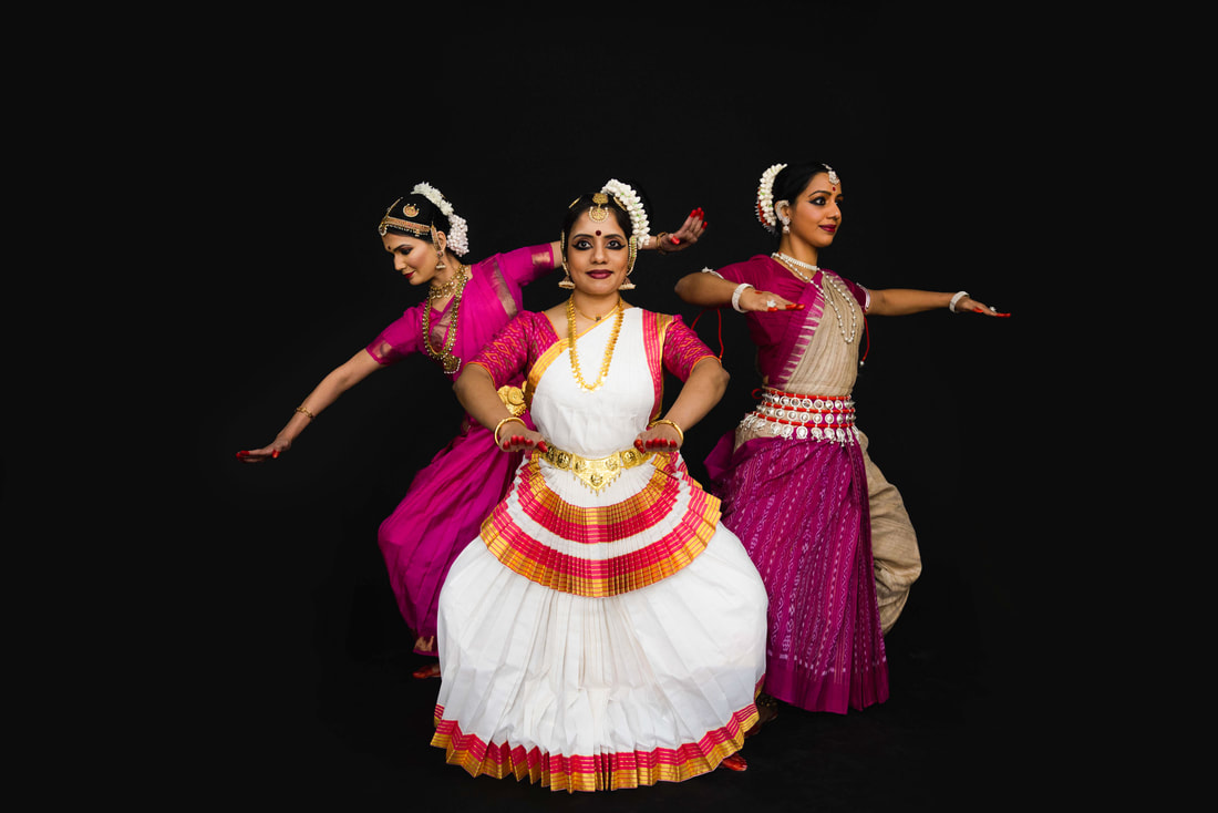 Three classical Indian dancers dressed in different types of traditional attire and ornamentation. One wearing white, pink and gold is standing in the front in a slight squat position with her arms held out in front of her torso. The other two are standing behind her facing opposite corners with their arms spread out to either side. Photo Credit: Olivia Moon Photography @halfasianlens