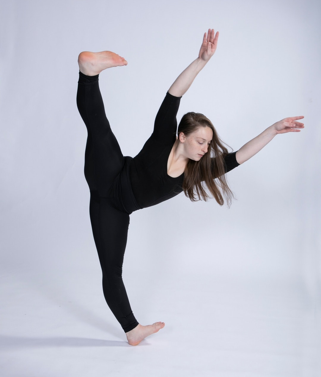 Woman with a long sleeve and full pant leotard stands on her left heel with her right leg extended