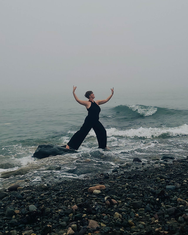 White woman in a black jumpsuit posing on a rock in the ocean with arms open above head
