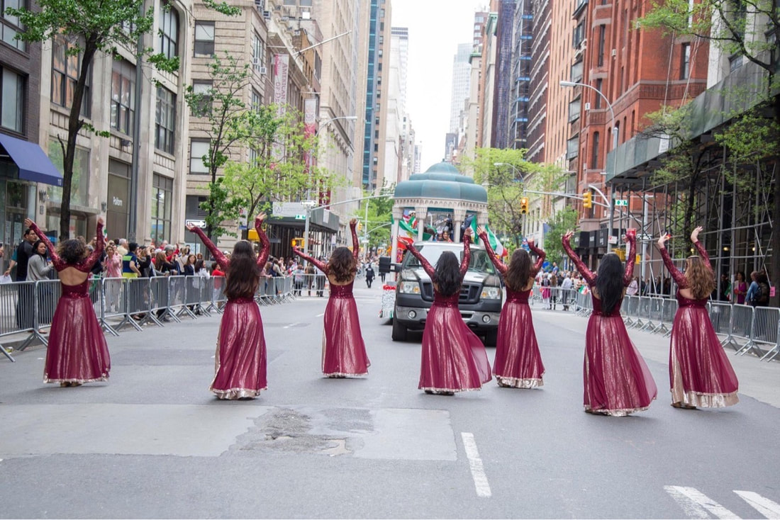 Seven dancers walk down a city street in a parade in long red dresses with their arms in the air
