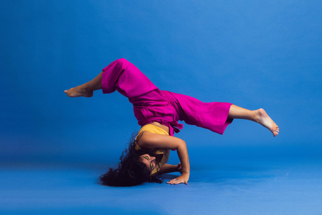 A dancer in front of a blue background wearing bright pink pants and an orange top. They are upside down with their head and arms braced on the floor as their legs are forming a split in the air above them with the front leg straight and back leg bent.