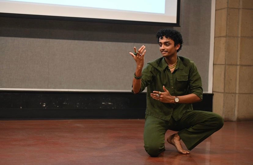 Brown skinned male wearing forest green long-sleeve shirt and matching bell-bottom pants is seated in a squatting position with one knee on the floor and hands close to the chest gesturing towards a kind, nurturing face. Background is concrete block with sound proofed board; floor is red concrete.