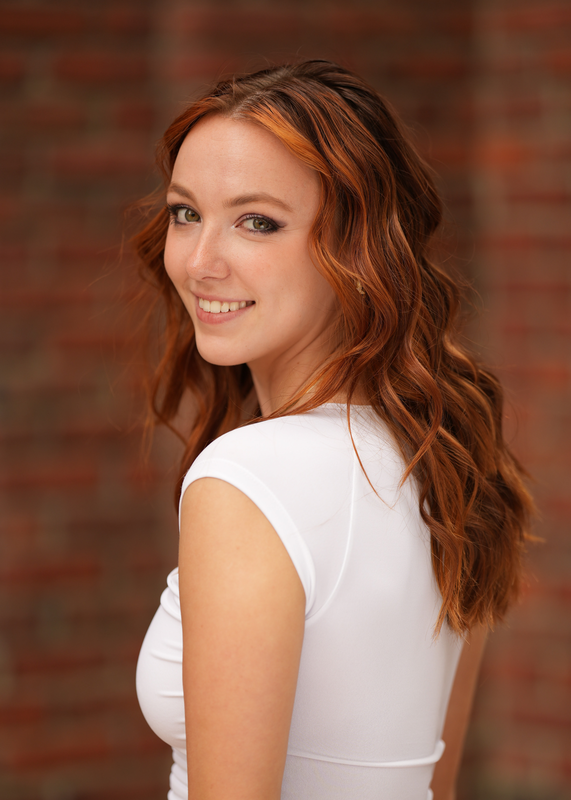 White woman (Annabelle Schultze) with curled copper hair wearing a white short sleeved shirt smiles over her shoulder