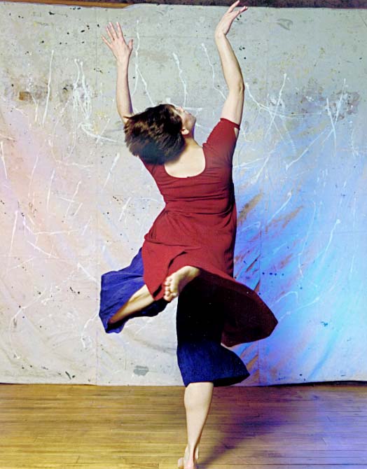 View of back of dancer with arms raised, one leg lifted, wearing red shirt and blue pants. 
