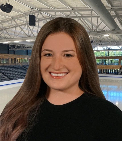Brooke is seen from the waist up with a big smile on her face as she looks at the camera. She has long brown hair and brown eyes and is wearing a black shirt. The background shows a skating rink, adding to the vibe of the picture.
