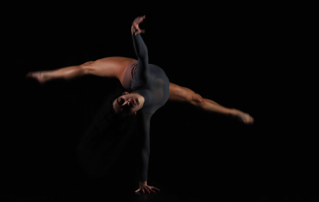 Carmen leaps in a dark space with one hand on the floor.