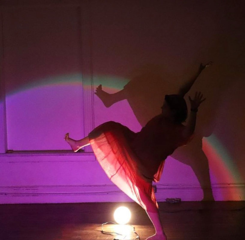 CJ stands at the center of a studio with beige wooden floors and white walls. They have their leg up as if stepping over a large whole. In between the step is a clamp light on the floor, which is shining at the camera. CJ is wearing a pink tulle dress, the bottom half of which is lit up by a purple hue off camera. Their hands are by their ears as if trying to balance. 