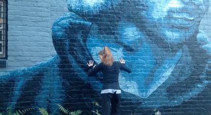 Person faces back Touches blue portrait on brick Looking to the ground