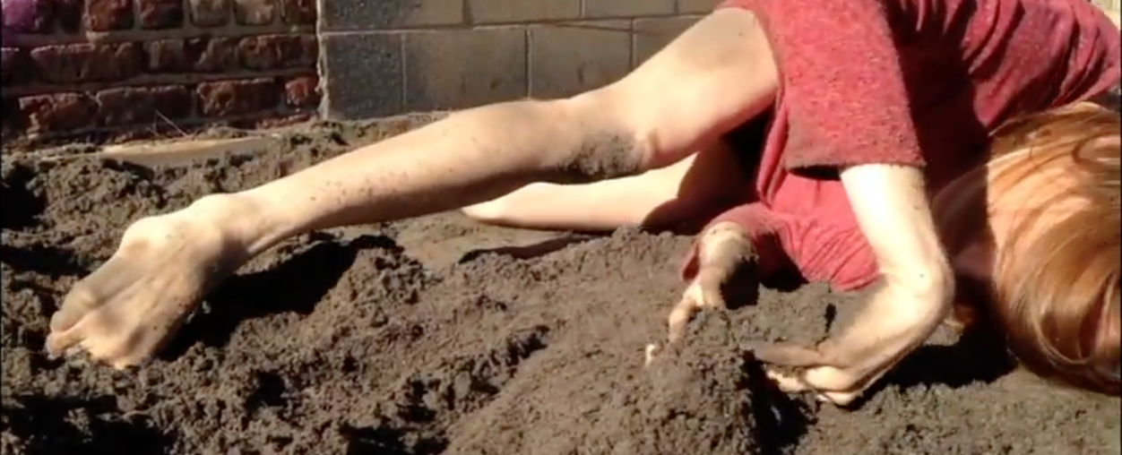 Dancer lies in sand  Gripping it in foot and hands Wearing a red shirt