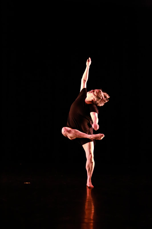 Dancer in all black room, throws one arm up, head to side, and one leg bent in front