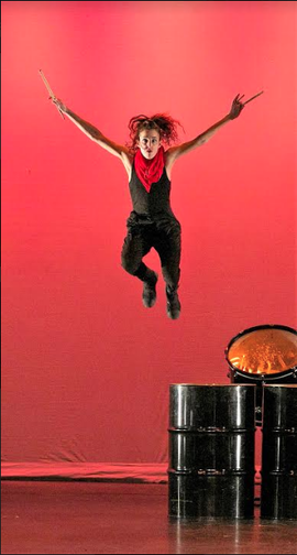 Woman in all black Jumps off drum holding drumsticks Red scarf on her neck