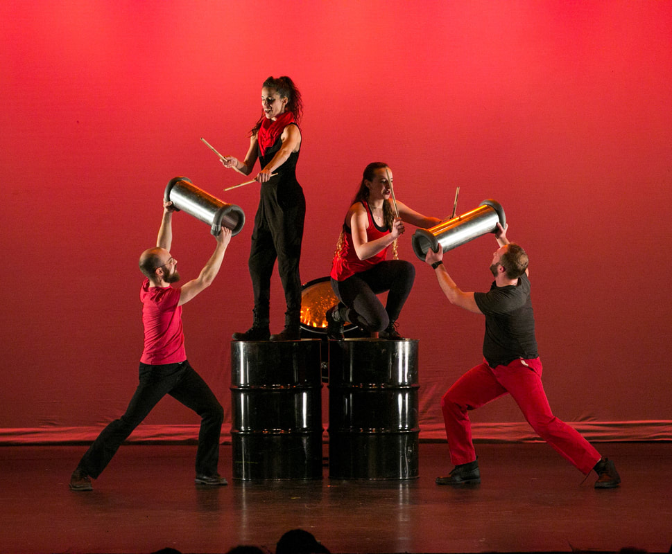 Four dancers on stage Two stand on large cylinders Two hold silver drums