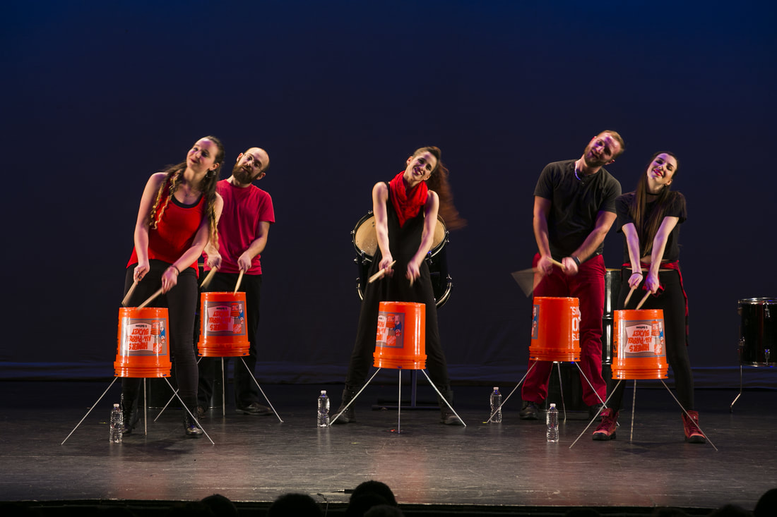 Five people on stage Playing orange buckets as drums Heads all tilting side