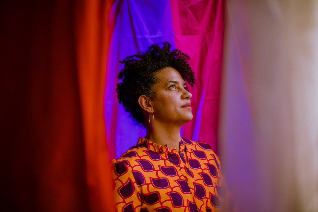 Jess, a brown skinned woman with curly hair, looks upward hopefully in 3/4 profile with a faint smile. She wears gold hoop earrings and bright geometrically patterned clothing in orange and purple, which relates to the vivid orange, purple, fuchsia and lavender fabrics which surround her foreground and background. 