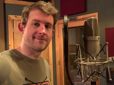 Ben Cuba, a white man, stands in a recording studio next to a microphone looking at camera with slight smile.
