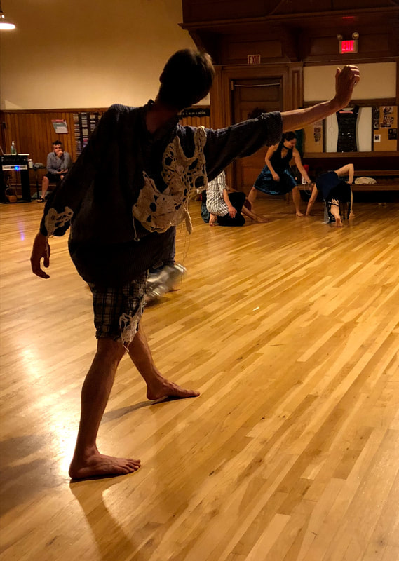 Back of a dancer One arm reaches out to side In room with wood floors