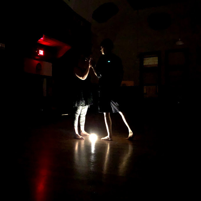 Two dancers stand close Lit up by one light below Everything is black