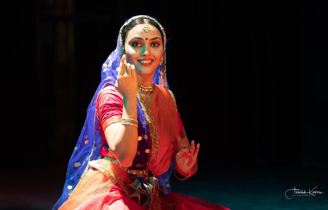 An image of a woman in bright pink blouse and skirt, and a blue dupatta (fabric) over her shoulder and around her head, wearing jewellery. The woman has a smile on her face, a bindi on her forehead, and hands in traditional dance pose