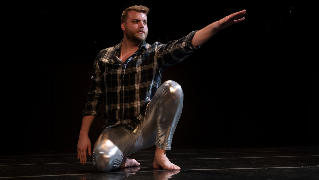 Bearded white man sits Wearing silver pants and plaid Reaching his arm out