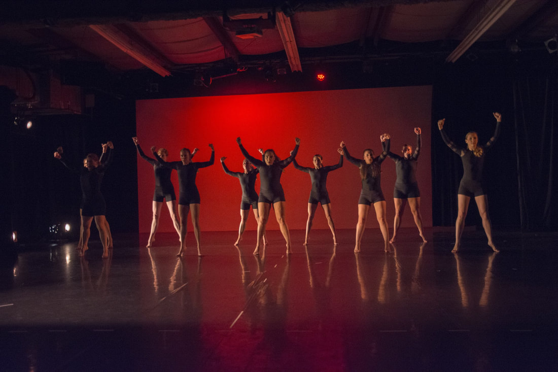 Nine dancers on stage, Up on toes, arms bent and raised, Black shorts, red background