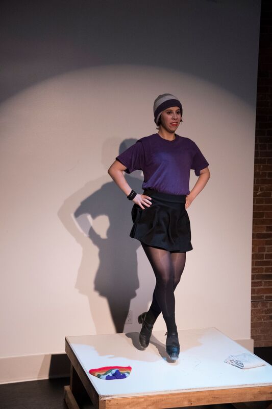 a white nonbinary person wearing an ace pride hat (grey, white, purple), a purple shirt, and a short black skirt. Their hands are on their hips and they are mid-air, feet pointing downwards.