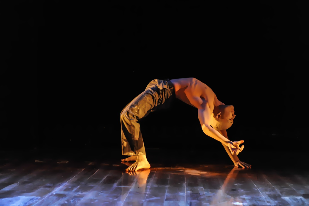 Dancer on a stage  Does backbend with one hand raised Wears no shirt, tan pants