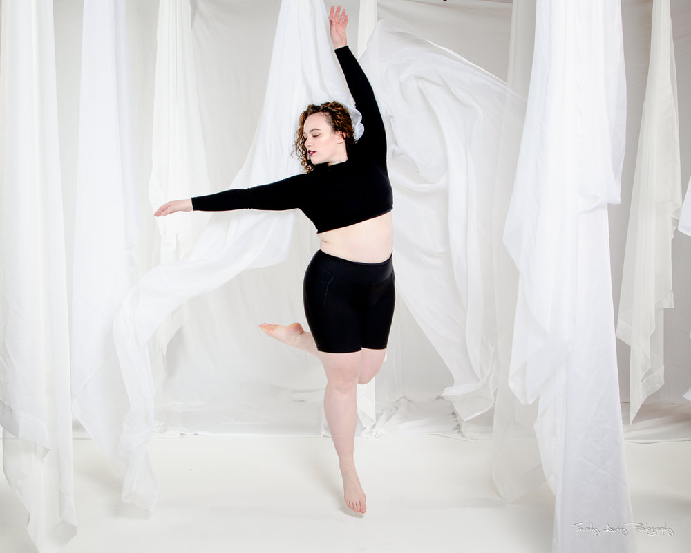 A woman in black shorts and a black long sleeved crop top jumps in white fabric with one leg swinging
