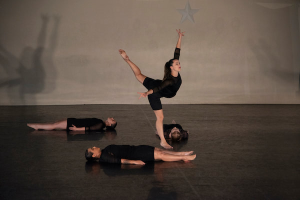 Four dancers on stage Three lie down on backs, one stands With arm and leg up