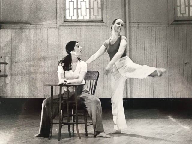 A woman sitting at a small table with another woman standing next to her with her leg lifted in the air