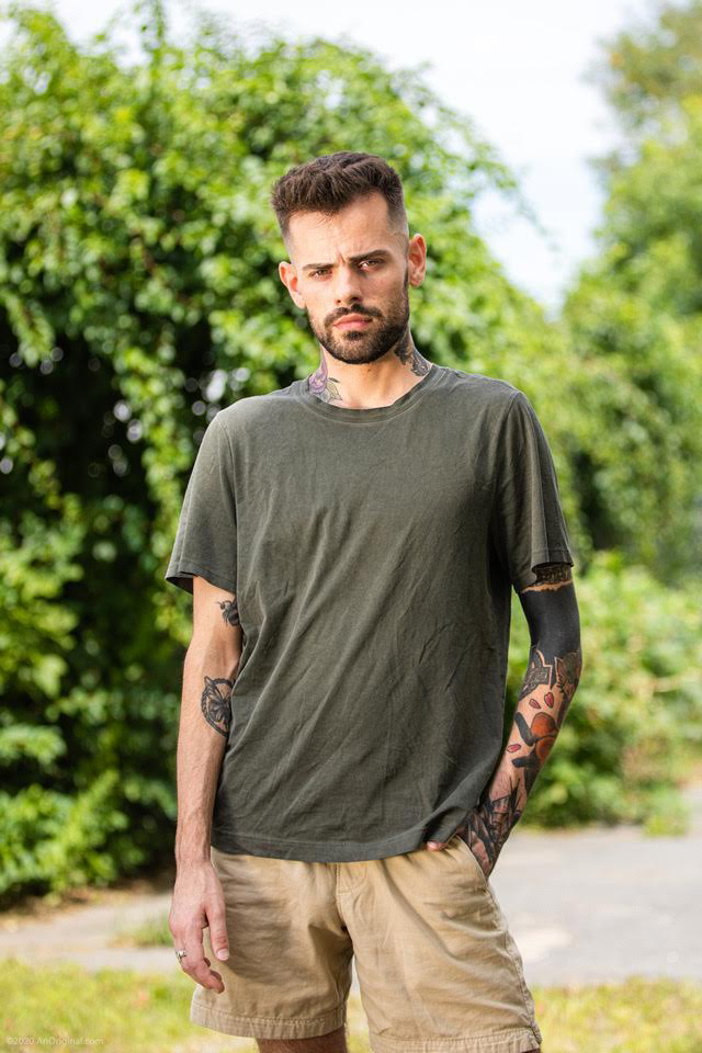 White man with tattoos Standing outside by bushes In shorts and a tee
