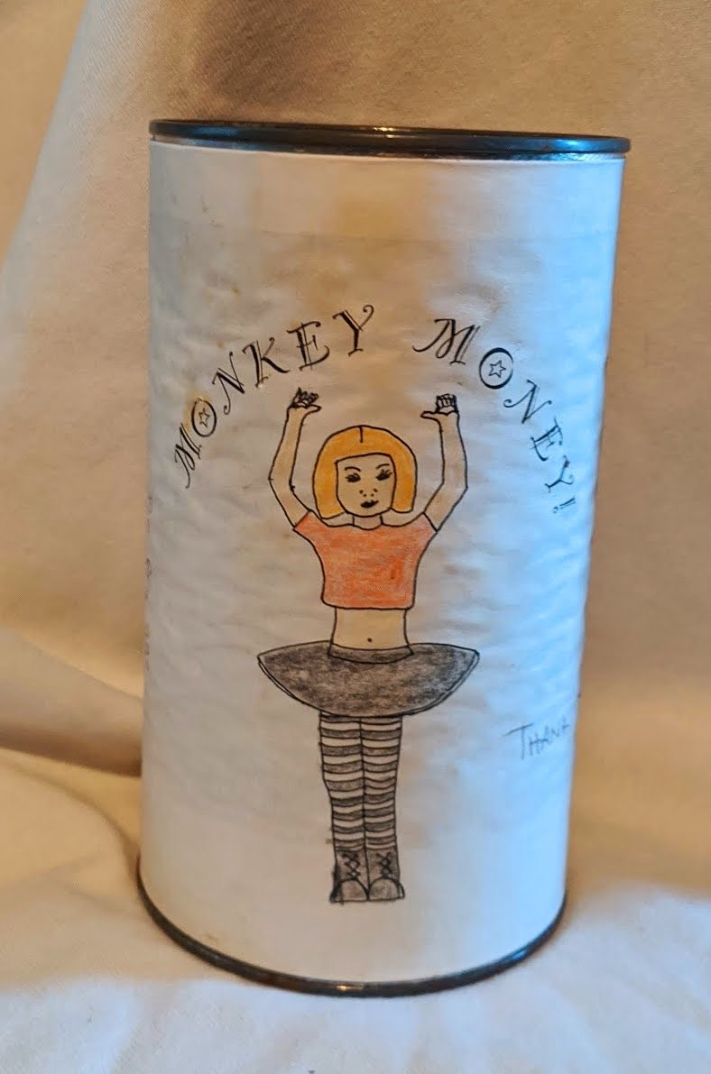 Coffee can covered in a white label. On the label is a drawing of a woman in an orange wig, black tutu and black and white striped tights with the text “Monkey Money”