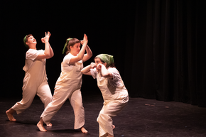 Three white women dance, Wearing white overalls and, Green bandanas too.