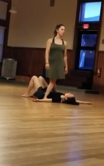 Two dancers rehearse, One lies on ground, one stands above, In room with wood floor