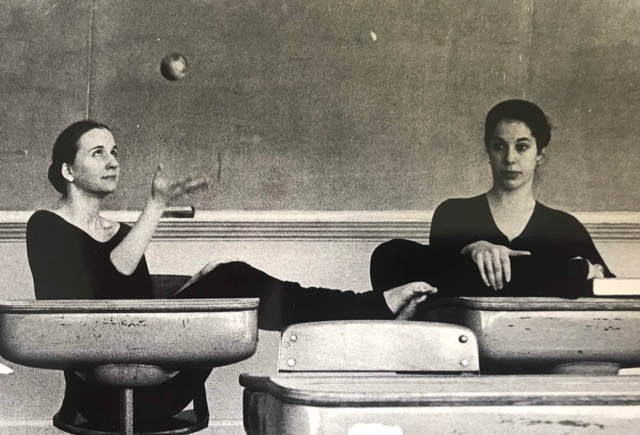 Two women at desks, Legs stretched on top, one throws ball Wear all black, hair back