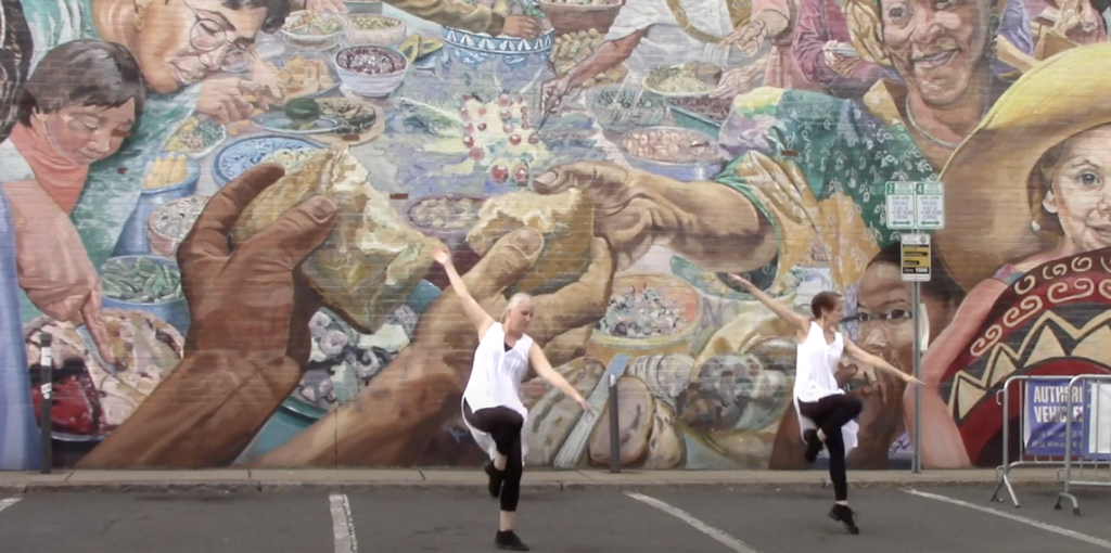 Two dancers pose in Front of mural of people Wear white tops, black pants