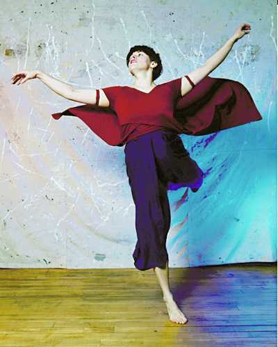 Short haired dancer in black pants and red shirt with cape throws arms, head, and one leg back.