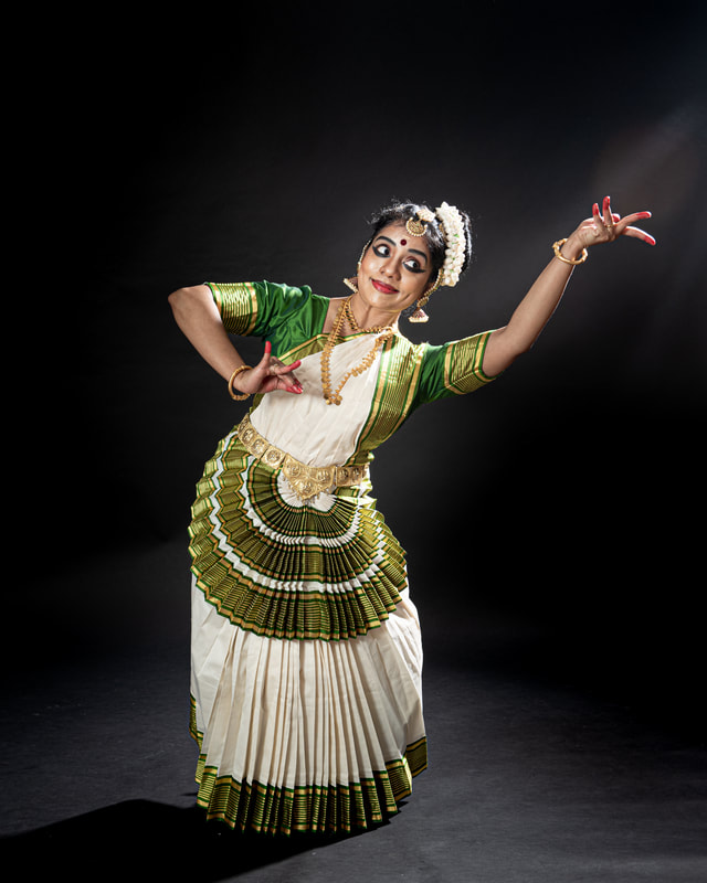 A full length image of dancer in white and green costume, gold jewelry and hair in a bun tied to the left .
