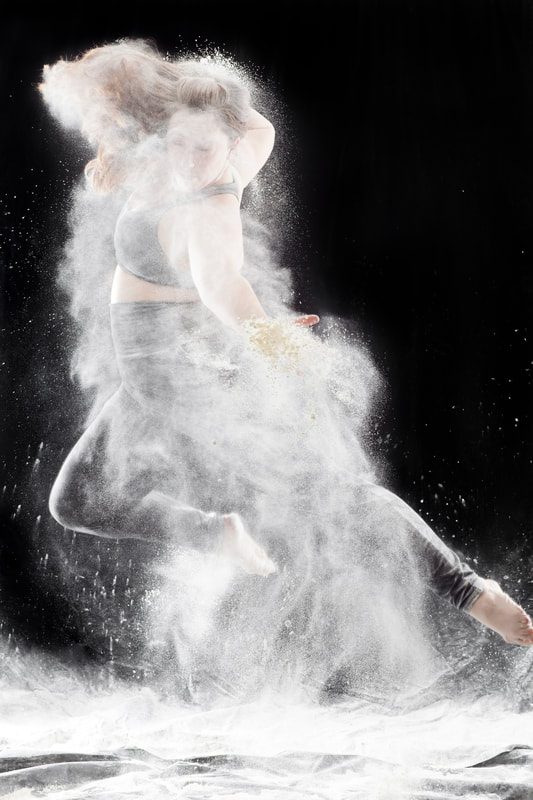Woman in black room Jumps, looking down, one leg bent White powder surrounds