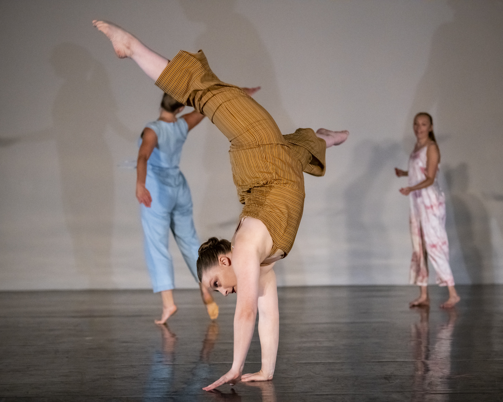 A woman in a yellow jumpsuit stands on her hands mid cartwheel