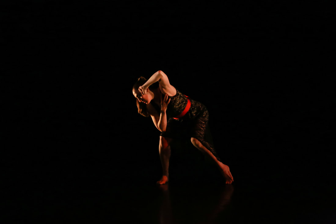 Dancer amid black  Bent knees, fist against her head Looking down at ground