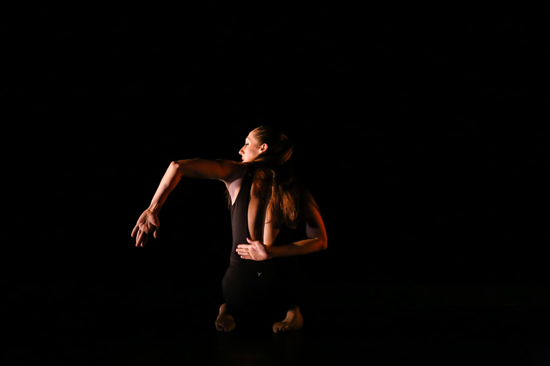 Dancer in black, one Arm bent, one reaches across  Her back, sits on knees 