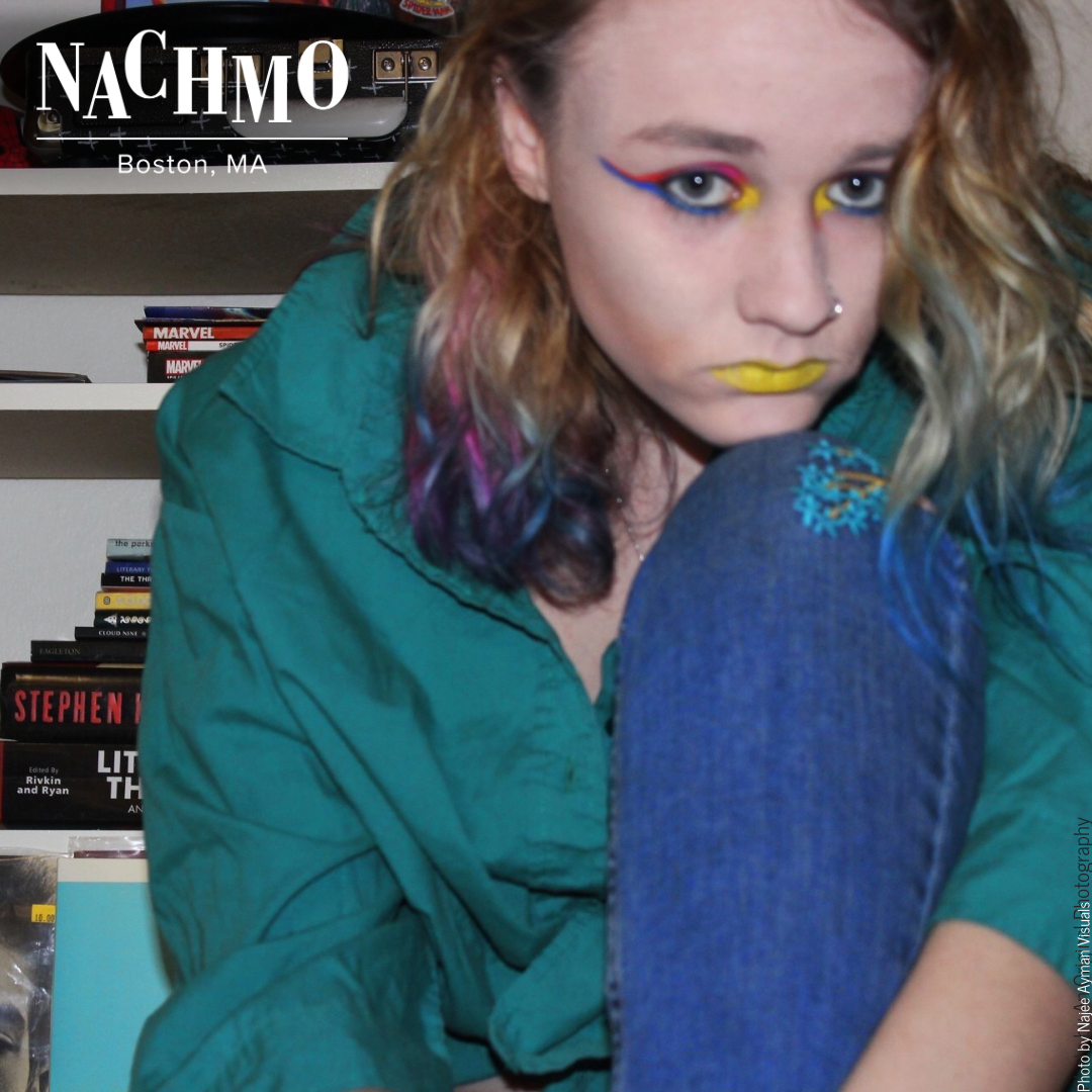 Blonde haired woman with blue, purple, and pink hair tips stares at the camera, chin on her knee. She wears pink, blue, and yellow eyeshadow with yellow lipstick. Behind her is a record player, comic books, and novels on a shelving unit.